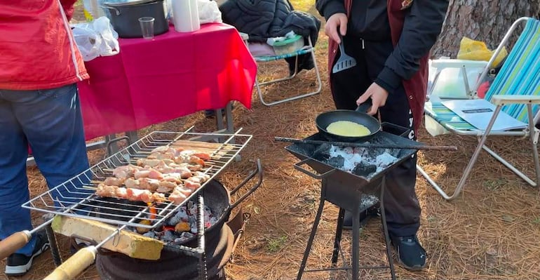 Dos hombres en un ambiente natural, uno con chaqueta roja y jeans, cocinando en parrilla con brochetas de carne.