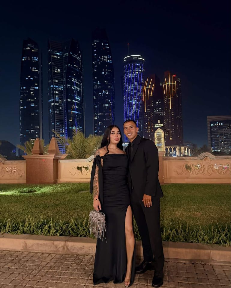 Pareja sonriente posando en un ambiente urbano nocturno. Ella lleva un vestido negro ajustado y él un traje negro.