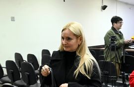 Ana Girala con abrigo negro y bolso, entrando a una sala vacía, mostrando expresión neutral.
