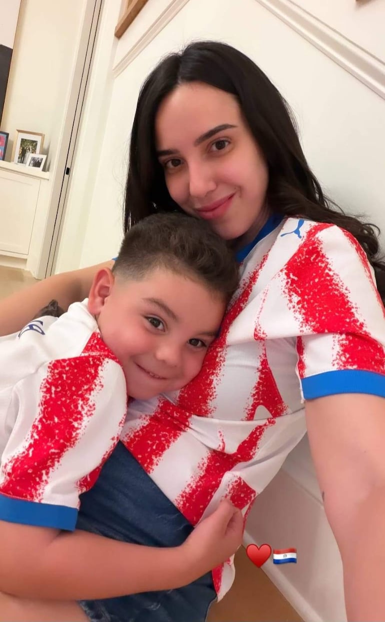 Mujer de pelo largo y oscuro abraza a un niño de cabello corto, ambos sonrientes con camisetas blancas y rojas de Paraguay.