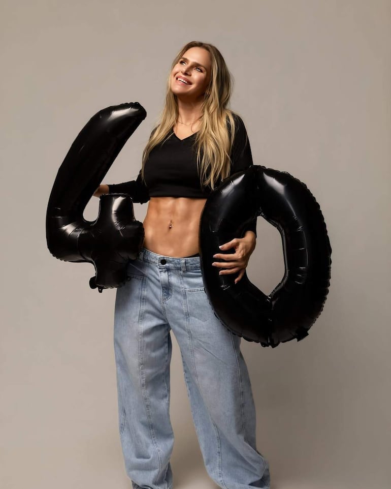 Mujer sonriente con cabello largo y suelto, viste camiseta negra y pantalones de mezclilla, sostiene globos de cumpleaños.