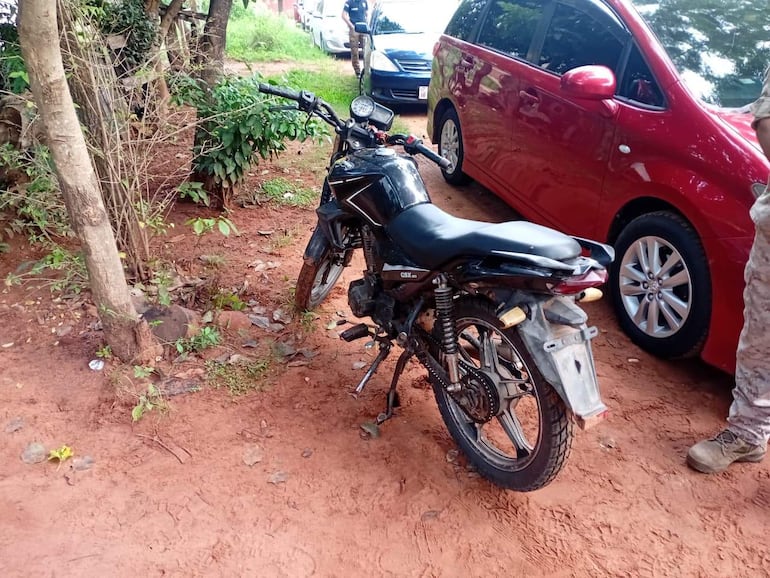 Motocicleta negra estacionada en suelo de tierra, rodeada de maleza y vehículos, sin personas visibles en el entorno.