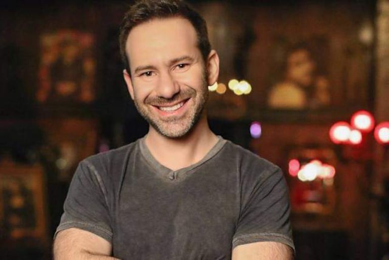 Leonid Radvinsky de pie con camiseta gris, sonriente, cruzando los brazos en un bar con luz tenue y decoración acogedora.
