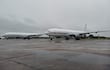 Dos aviones Boeing 767 blancos en un aeropuerto con cielo nublado y pavimento húmedo, sin personas visibles.
