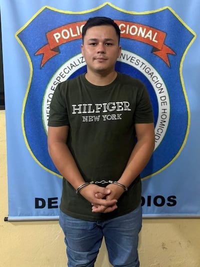 Joven de pie con camiseta verde y manos esposadas, frente al escudo de la Policía Nacional en fondo azul.