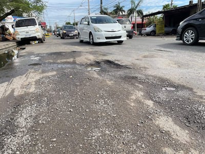 Avenida Santa Teresa con baches y baches visibles, vehículos de diversos colores en el fondo y un comercio de copias de llaves al lado.