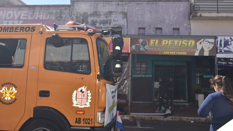 Camión de bomberos naranja con dos hombres, uno en uniforme, evaluando el incendio en El Petiso, una tienda de bazar.