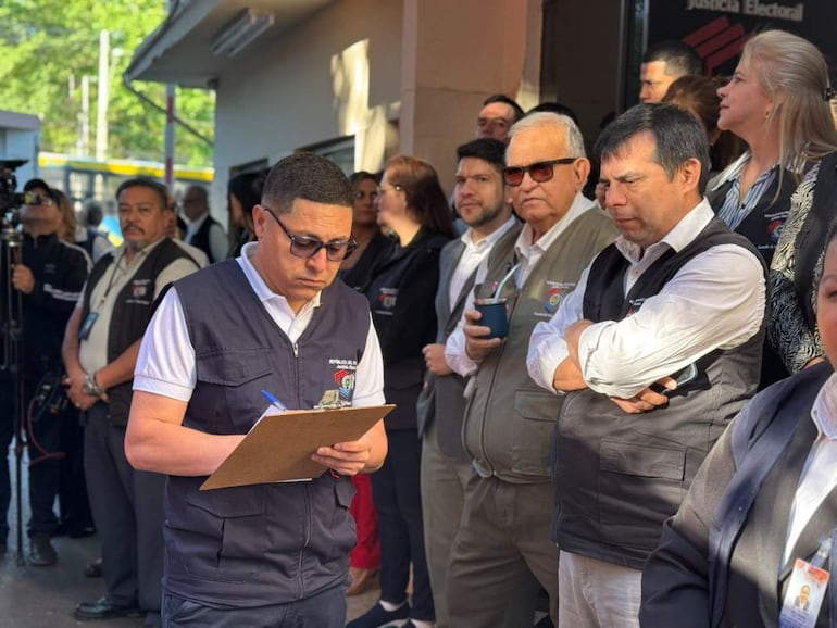 Hombres y mujeres en chaquetas formales, algunos con notas, cerca del edificio de la Justicia Electoral, con serias expresiones.
