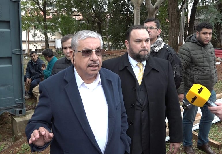 Dos hombres de pie con trajes oscuros, uno hablando y otro escuchando; cuatro hombres adicionales en ambiente de evento informativo.