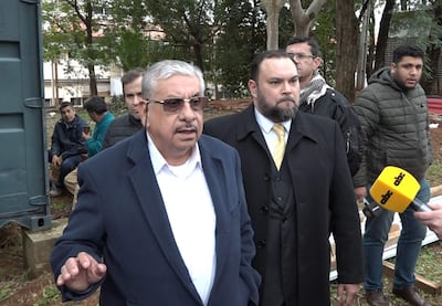 Dos hombres de pie con trajes oscuros, uno hablando y otro escuchando; cuatro hombres adicionales en ambiente de evento informativo.