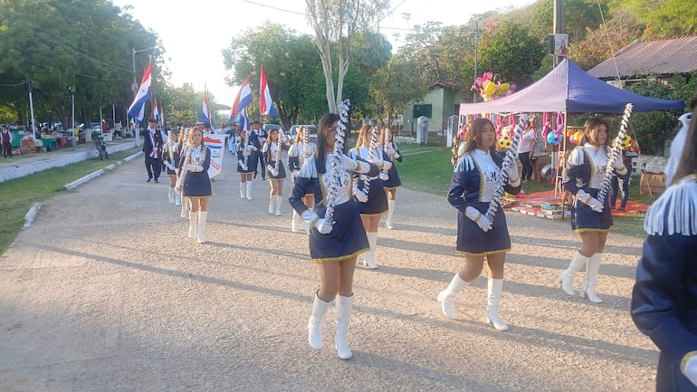 Grupo de estudiantes en trajes azules marchan con banderas en Fuerte Olimpo, bajo un ambiente festivo entre árboles y un mercado.