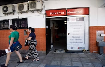 Dos mujeres, una con camiseta verde y la otra con camiseta oscura, caminan juntas conversando frente a un policlínico municipal.