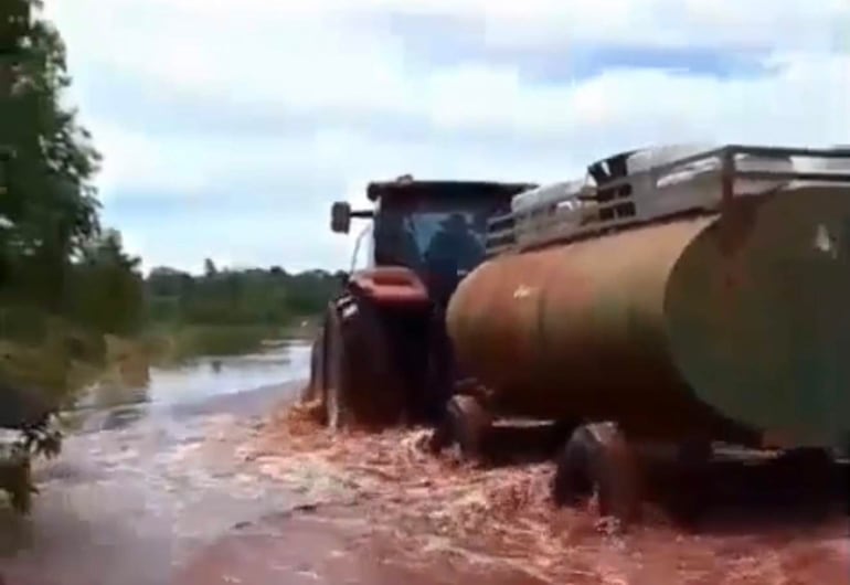 Un tractor rojo se desplaza por una ruta inundada rodeada de vegetación en un entorno agrícola, generando ondas de agua.