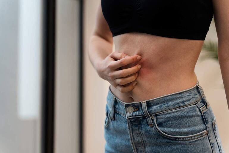 Mujer con piel clara, sosteniendo su abdomen, con sostén negro y pantalón de mezclilla claro, en un entorno interior.