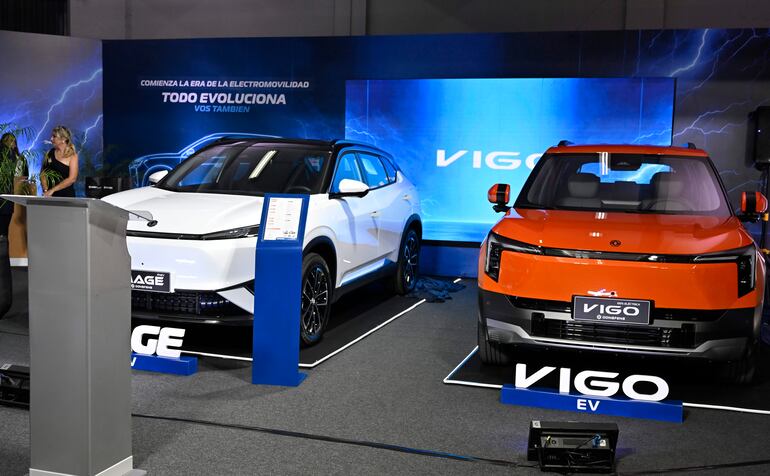 Mujer de pie junto a autos eléctricos blanco y naranja, con pantalla que muestra 'COMIENZA LA ERA DE LA ELECTROMOVILIDAD'.