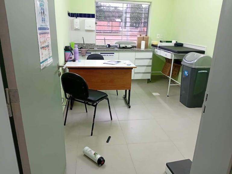 Consultorio vacío con escritorio y sillas. Documentos y utensilios médicos en mesada, luz natural entra por la ventana.