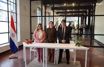 Natalia Fuster en blusa blanca y pantalón rosa, junto a Ortiz Semidei y Gayoso, todos de pie en un ambiente formal con la bandera de Paraguay.