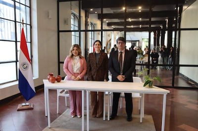 Natalia Fuster en blusa blanca y pantalón rosa, junto a Ortiz Semidei y Gayoso, todos de pie en un ambiente formal con la bandera de Paraguay.