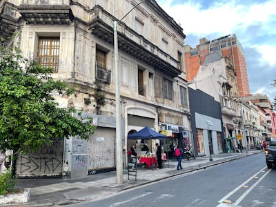 Persona vestida de negro en un puesto de venta, con calles desgastadas y árboles alrededor en un ambiente urbano.