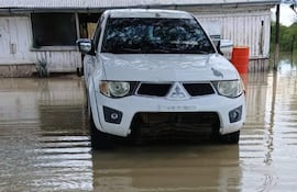 Vehículo SUV blanco sumergido en agua, con una casa de paredes desgastadas y un barril anaranjado al fondo.
