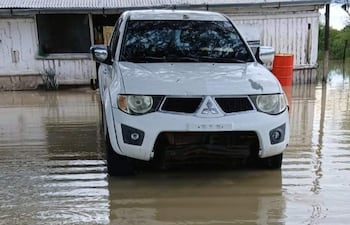 Vehículo SUV blanco sumergido en agua, con una casa de paredes desgastadas y un barril anaranjado al fondo.