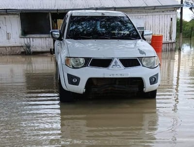 Vehículo SUV blanco sumergido en agua, con una casa de paredes desgastadas y un barril anaranjado al fondo.