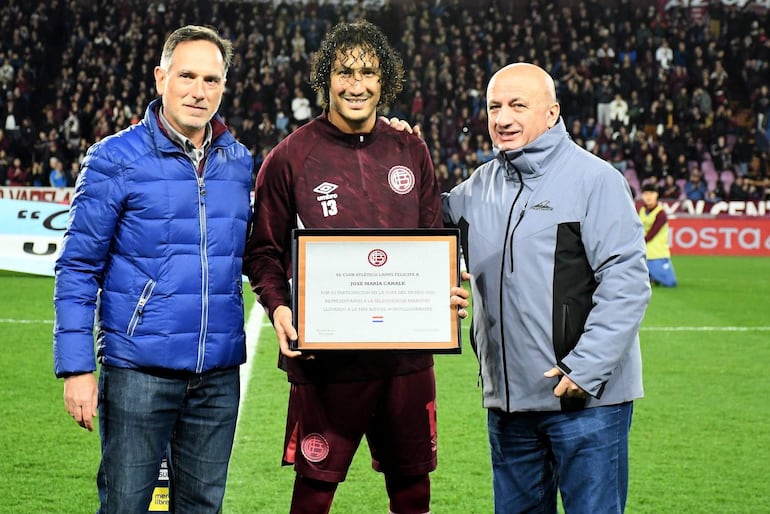 José María, con camiseta número 13, sostiene un certificado, mientras dos hombres con abrigos lo acompañan en un estadio lleno.