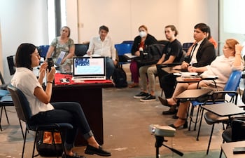 Mujer de cabello oscuro y corto en camisa blanca habla con micrófono, rodeada de oyentes tomando notas en un aula.