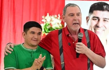 Hombre canoso con camiseta roja sostiene micrófono y habla, mientras joven aplaude en un ambiente festivo de fondo rojo.
