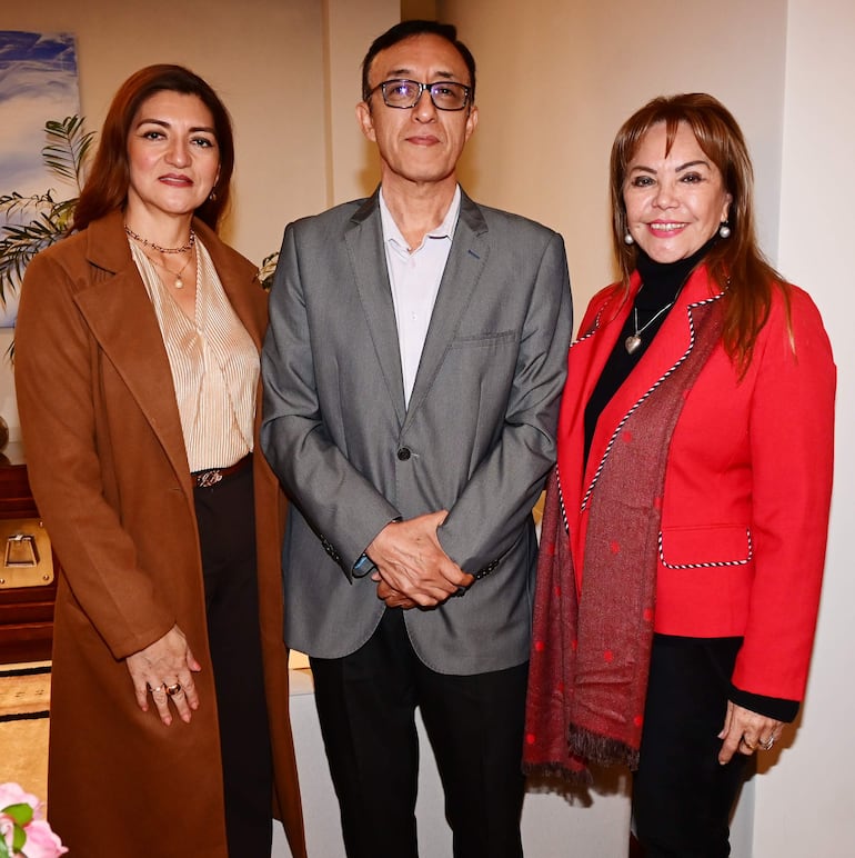 Mujer con abrigo marrón, hombre en saco gris y mujer con chaqueta roja sonríen juntos en un entorno decorativo.