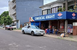 Hombre sentado frente a 'Comercial Anahy', rodeado de vehículos en un barrio residencial, con árboles y elementos publicitarios visibles.