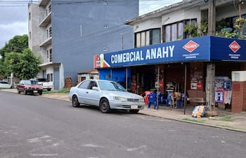 Hombre sentado frente a 'Comercial Anahy', rodeado de vehículos en un barrio residencial, con árboles y elementos publicitarios visibles.