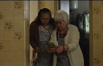 Katty Pacuá con abrigo marrón y Amada Gómez en suéter blanco, observan un brócoli en una cocina decorada con azulejos de flores.