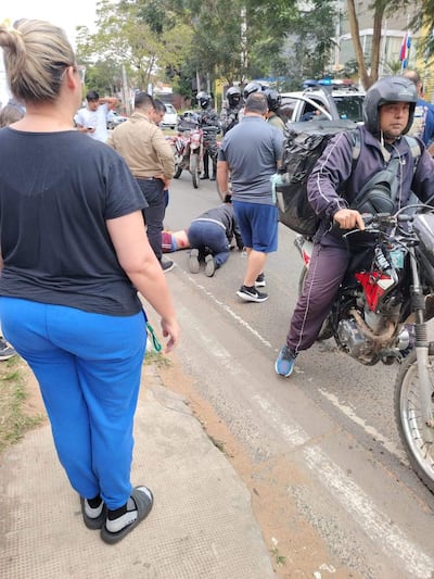 Hombre tendido en el suelo con herida en la pierna, rodeado por testigos y un motociclista con casco atento a la situación.