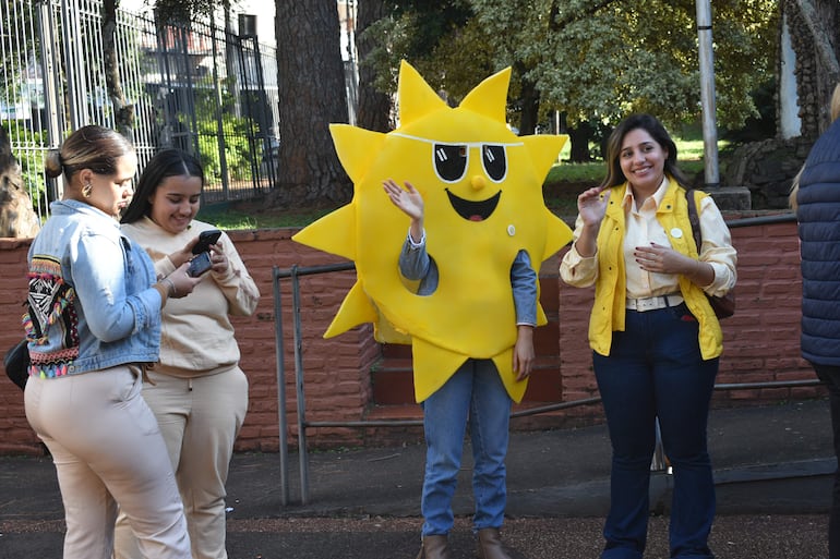 Mujeres conversan en un parque; una en disfraz de sol saluda, otra sonríe con chaleco amarillo. Ambiente alegre.