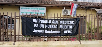 Fachada de la Casa del Maestro con un gran cartel negro que dice 'UN PUEBLO SIN MÉDICOS ES UN PUEBLO MUERTO' y una bandera paraguaya.