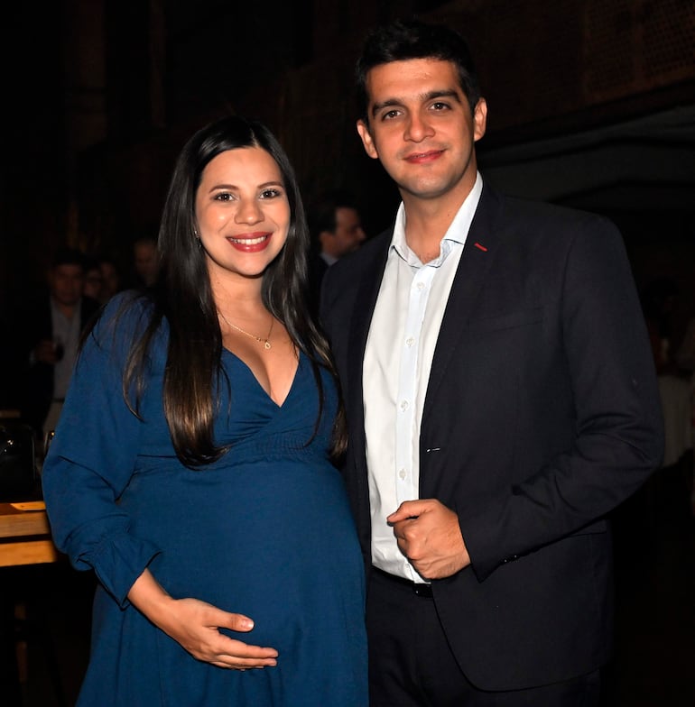 Mujer embarazada en vestido azul sonríe sosteniendo su vientre, junto a un hombre con camiseta blanca y saco oscuro en evento social.