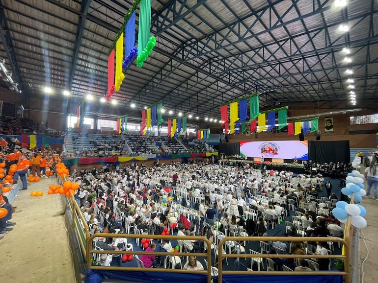 Gran multitud de personas sentadas en filas, vestidas de blanco, en ambiente festivo con globos y banderas de colores.