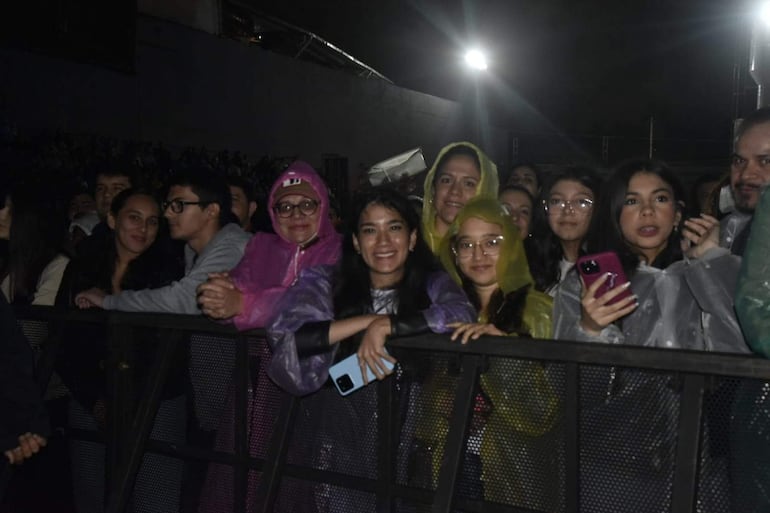 Grupo de jóvenes con impermeables de colores sonríen y esperan el show de Tan Biónica en medio de la lluvia.
