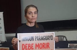 Mujer con cabello largo y lacio, camiseta negra y jeans, sostiene un cartel con mensaje de protesta en un salón.