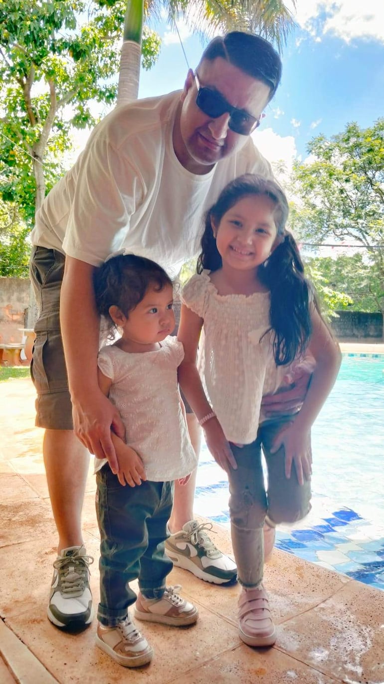 Hombre de pie con gafas de sol, junto a dos niñas sonriendo, en un entorno natural junto a una piscina.