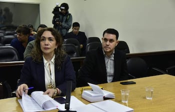 Hernán Rivas en camisa a rayas y saco negro, junto a Alba Zaracho con blazer oscuro, en una sala con documentos sobre la mesa.