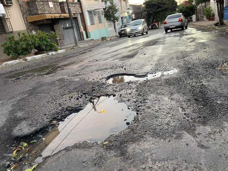 Calle urbana deteriorada con baches y agua estancada, vehículos en movimiento entre edificaciones residenciales y comerciales.
