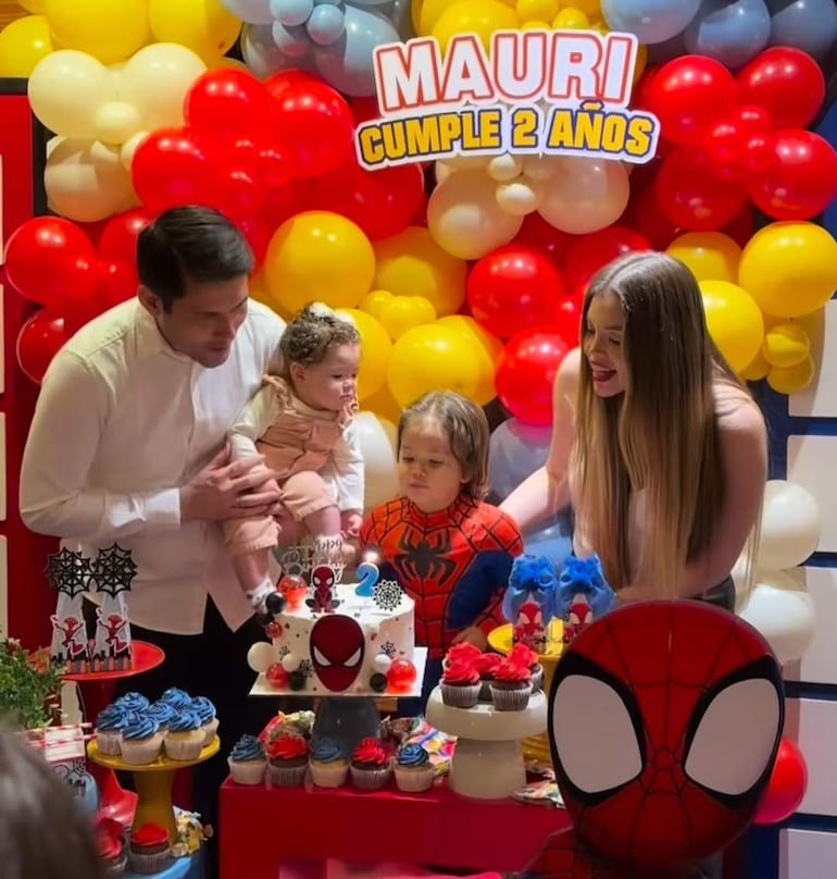 Un niño con camiseta de Spider-Man sopla las velas de un pastel, rodeado de adultos emocionados y decoración colorida.