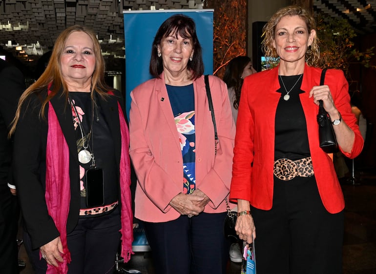 Tres mujeres sonrientes en evento: una con blusa negra y chal rojo, otra con blusa azul y chaqueta rosa, y la última con blusa negra y chaqueta roja.