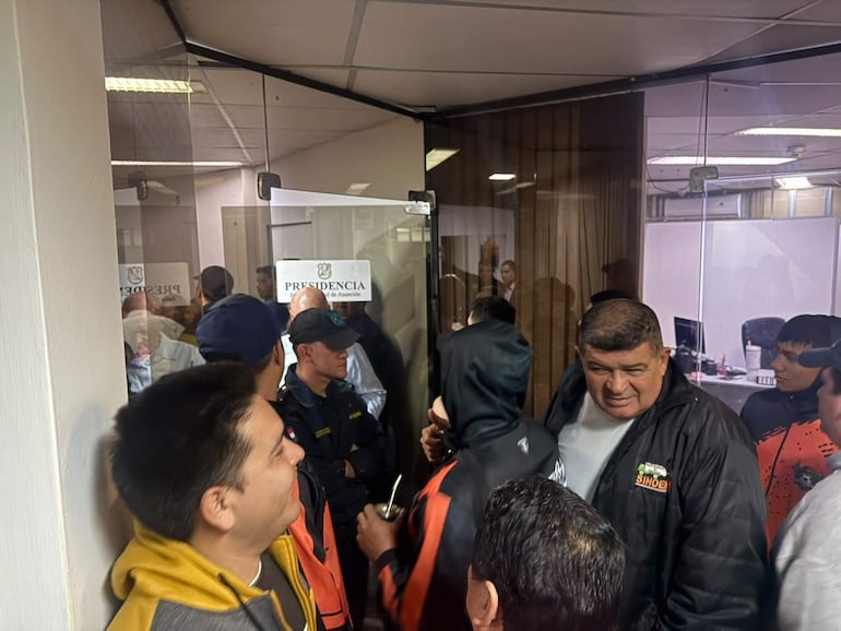 Carlos Ferreira Bogado con chaqueta amarilla y camiseta blanca, junto a otros hombres en chaquetas oscuras, conversando en una oficina.