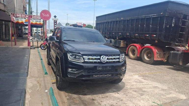 Un vehículo SUV negro estacionado en un área comercial, con comercios visibles en el fondo y un camión de carga.