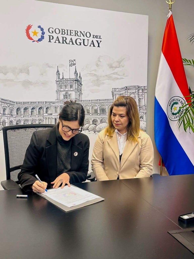 Adriana Ortiz, con chaqueta negra y gafas, firma documentos, mientras Natalia Fuster Cascio, con blusa blanca y abrigo beige, observa atentamente.