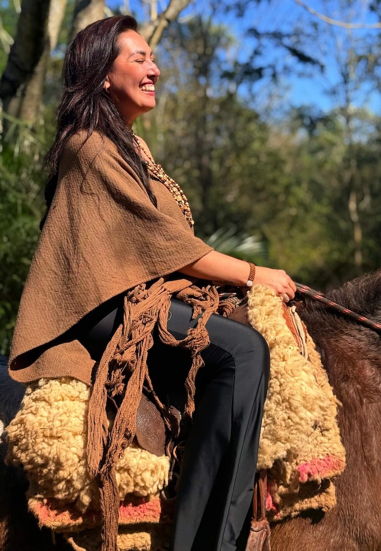 Mujer con poncho marrón y collar de cuentas, sonríe montando un caballo en un paisaje natural.