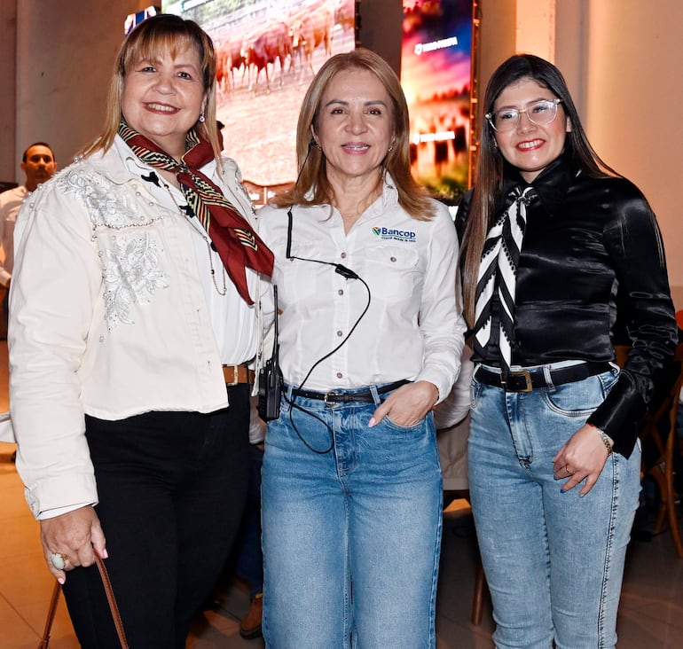 Tres mujeres sonrientes: una con bufanda marrón, otra con micrófono y logo de Bancop, y la última con gafas y blusa negra con lazo blanco.